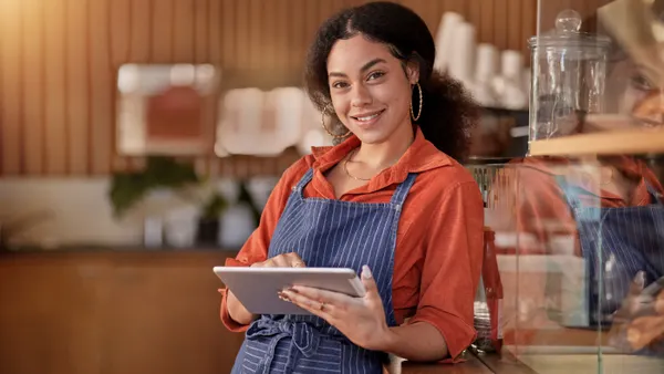 Lady with large earrings in orange shirt and blue striped apron holding a tablet
