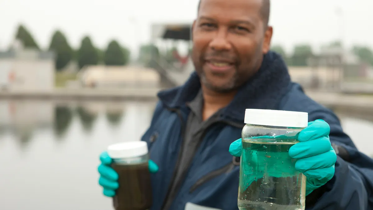 Man smiling at the camera with medical gloves holding up a jar of murky water in front of water facility