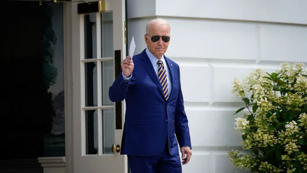 An older White man in dark aviator sunglasses and a suit gives someone out of frame a genial hand gesture.