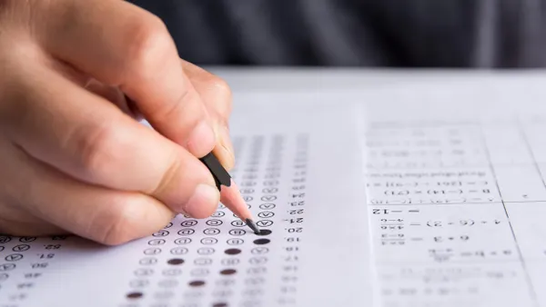 A close-up of a hand holding a pencil, filling in answer bubbles on a standardized test sheet. The page also includes math equations in a grid format. The person’s torso is slightly blurred in the background.