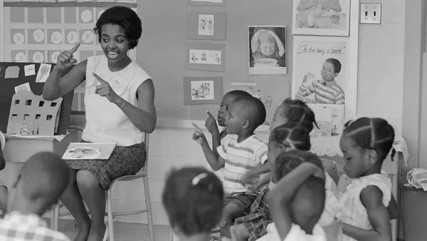 An adult is sitting in a chair at the front of a classroom and pointing in the air. Young children are seated on the floor around the adult and some are also pointing.