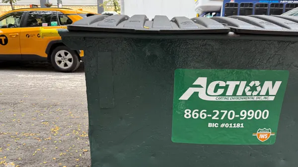 A commercial dumpster sits on the sidewalk next to a busy avenue with a taxi. The dumpster bears the "Action Environmental" logo.
