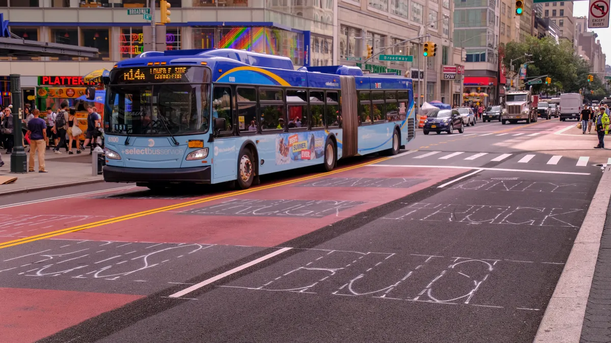 A New York city articulated transit bus follows a bus lane marked in red, passing people on a sidewalk.