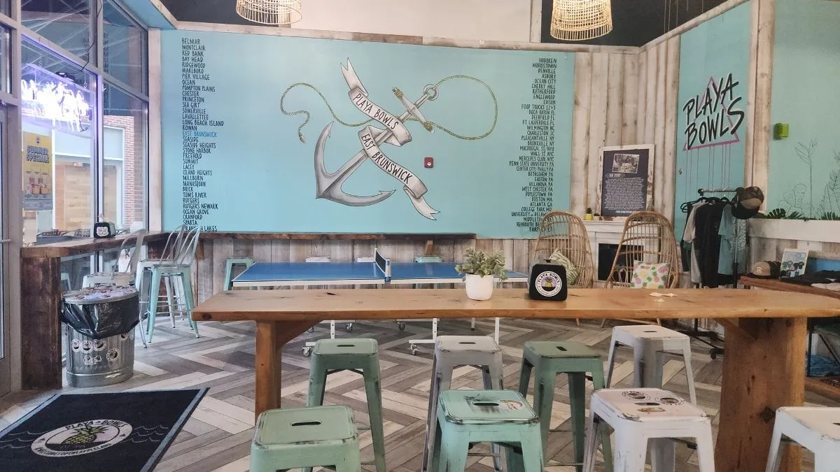 An image of a Playa Bowls with a table and stools. There is a ping pong table in the background and a wall with an anchor that says East Brunswick.