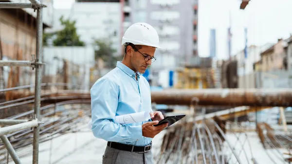 A person in a button down blue shirt holds a tablet while wearing a white hard hat on a jobsite.