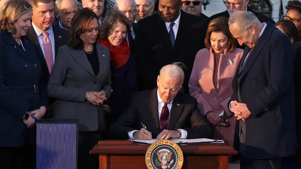 President Joe Biden seated at a table with pen in hand surrounded by men and woman officials.