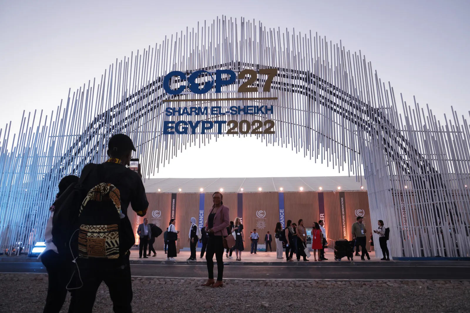 People walk toward a metal archway with vertical rods with a sign reading "COP27 Sharm El-Sheikh Egypt 2022" at the middle. A large building is on the other side.