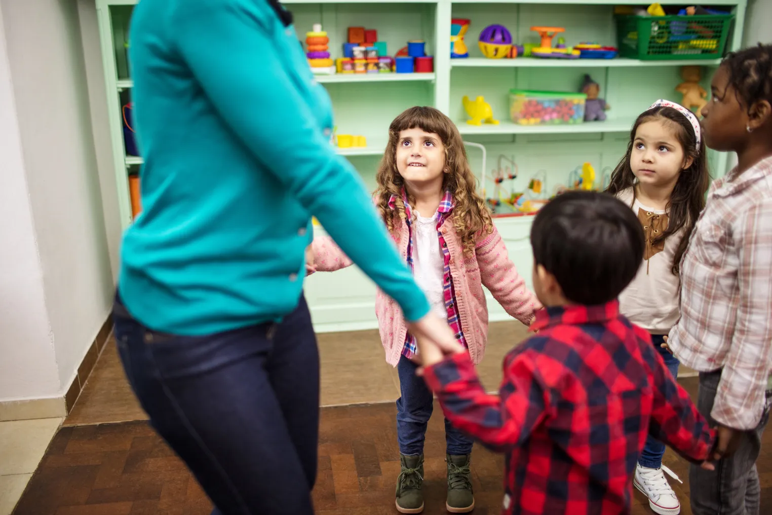 An adult stands in a circle with young children in a classroom. They all are holding hands.