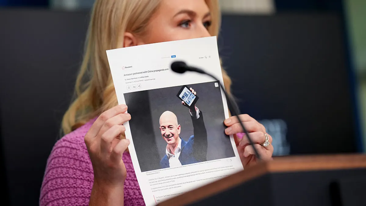 A person with long blond hair, wearing a pink shirt, holds up a photo of another person.