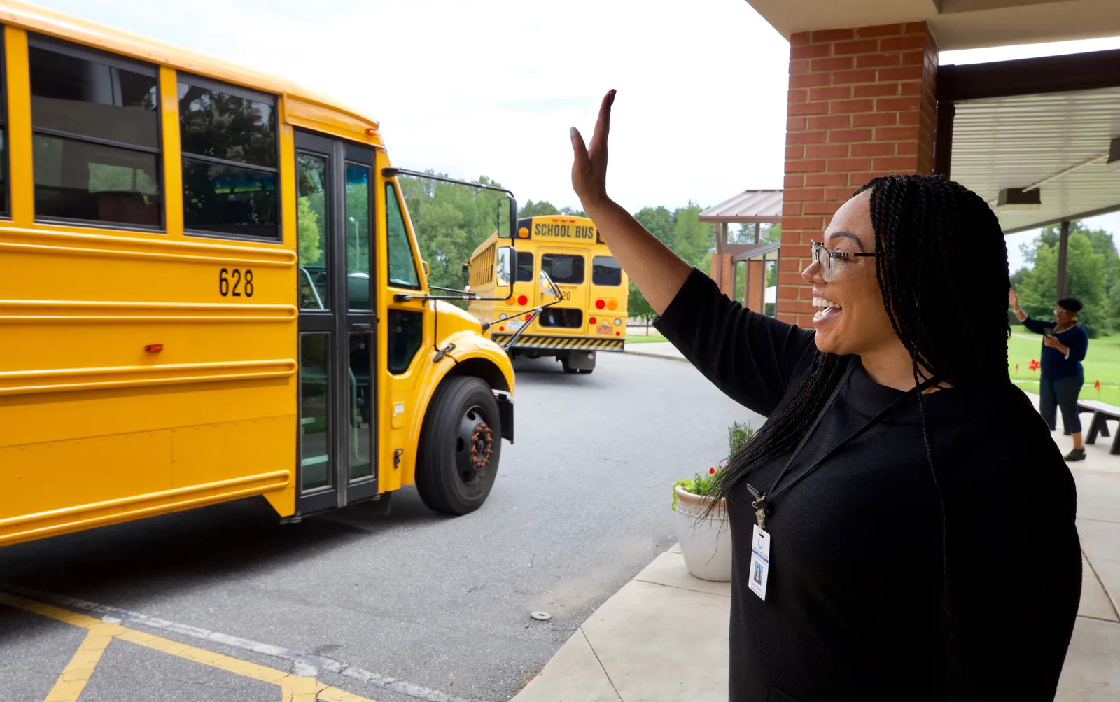 An adult stands outside a school building with arm up in arm. Person is facing two yellow school buses.