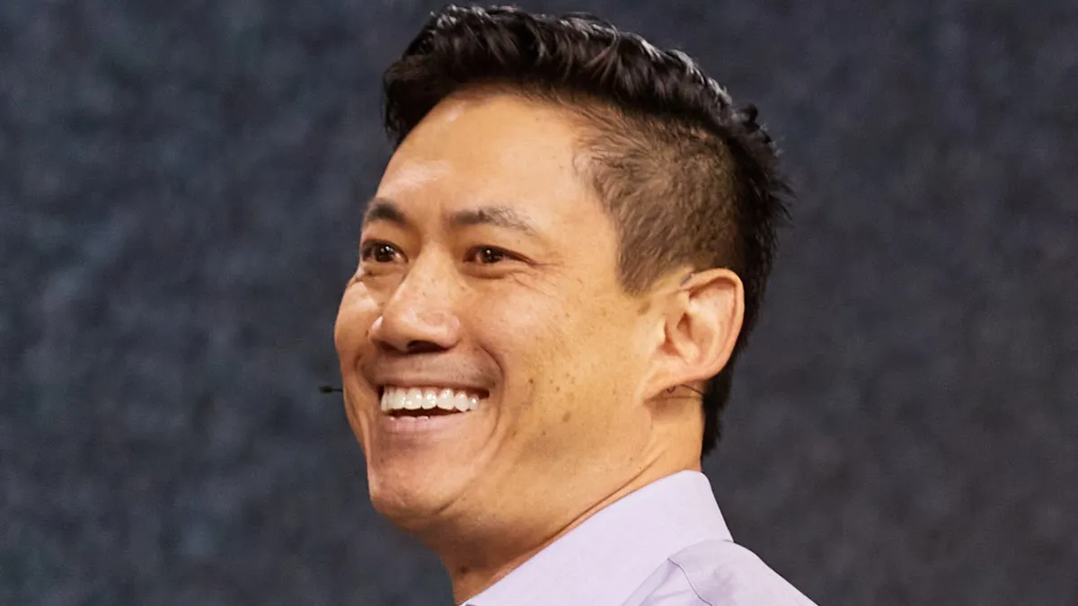 An image of a man smiling and looking off to the side. He has a headset microphone on his right ear.