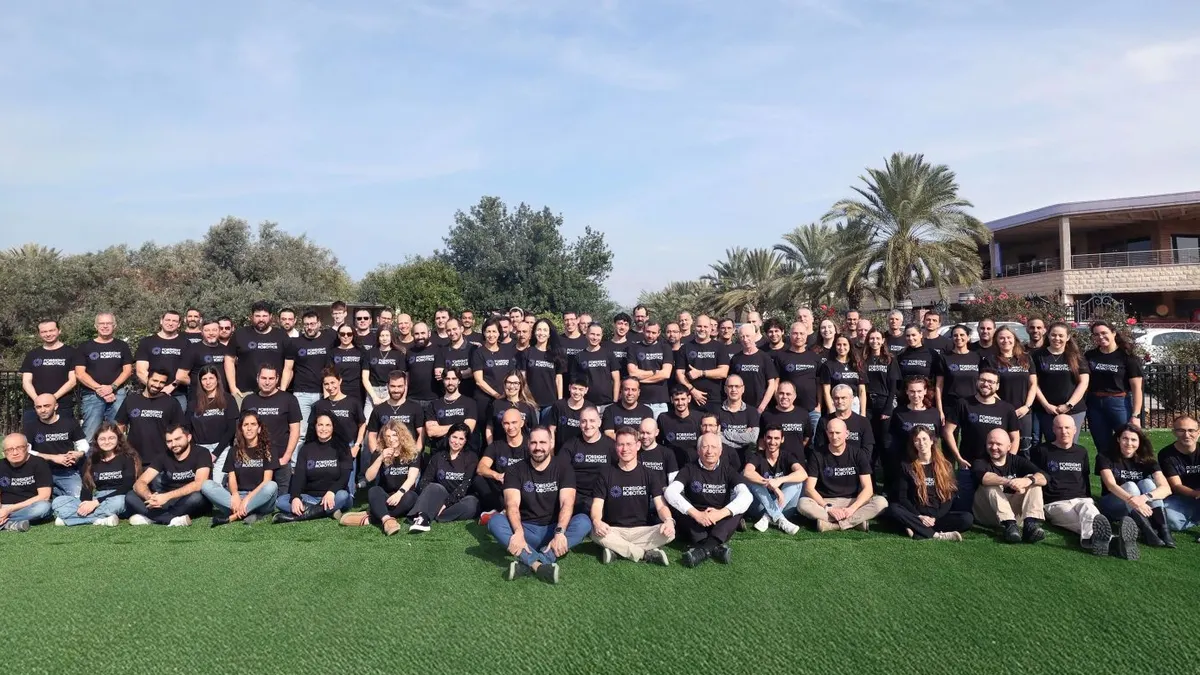 A large group of ForSight Robotics employees wearing company T-shirts sits on the lawn outside of their office.