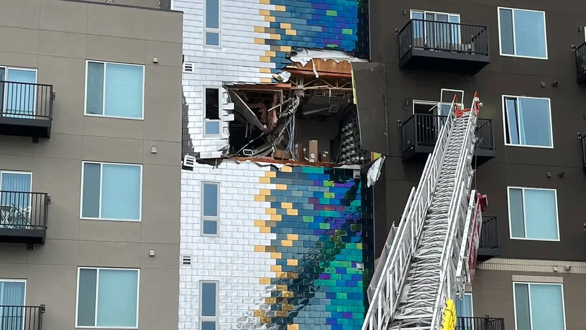 An apartment building with a blue brick wall with a large hole in it. A fire truck crane is next to the hole.