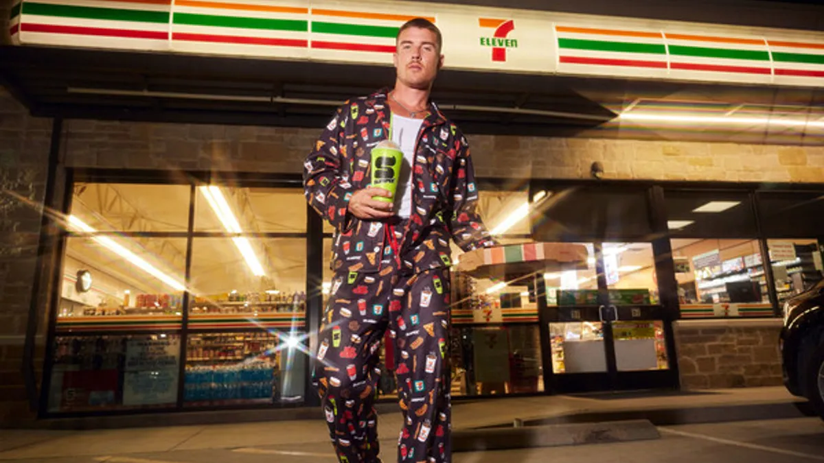 A model wearing black, red and green 7-Eleven holiday pajamas in front of a 7-Eleven holding a Slurpee drink