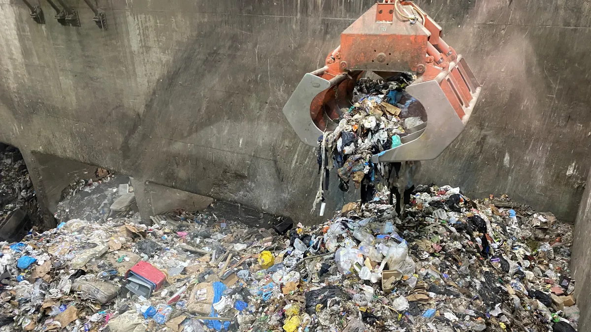 A large crane drops mixed trash into a large concrete pit in an enclosed building.