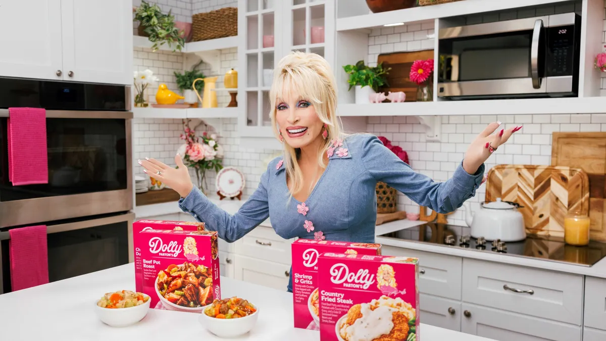 Dolly Parton stands in a kitchen next to her new line of frozen meals