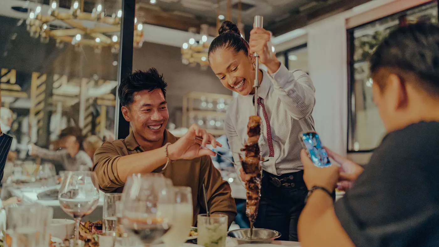 A person in a white shirt hold up a stick of meat while a person at a table takes a portion of meat off the stick. Another person is taking a photograph with a smartphone