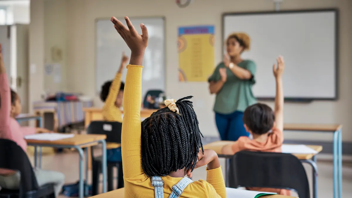 Elementary school students raise their hands as they sit at their desks in a classroom with their teacher asking a question in front of them.