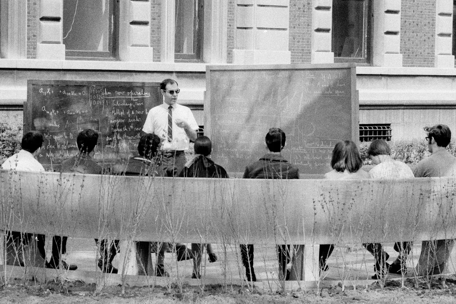 A professor standing between two blackboards lectures a group of students sitting on a large bench.