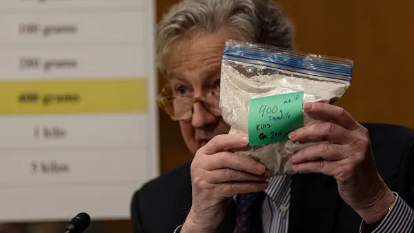 A person sitting in a wood-panel room holds up a clear plastic bag with white powder. The person's face is partially hidden by the bag.