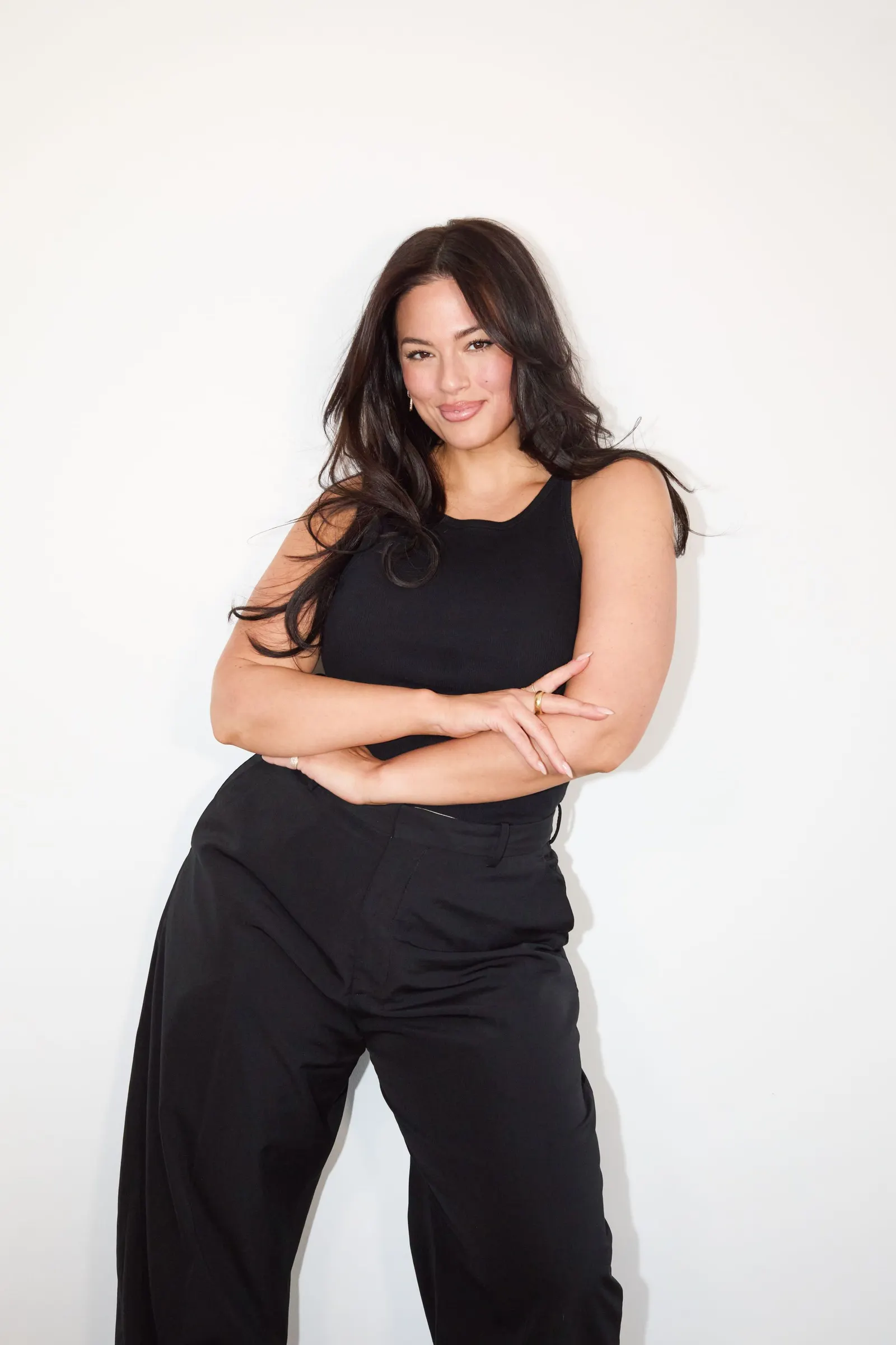 A woman in a black sleeveless pants look stands facing the camera with her arms crossed and smiling.