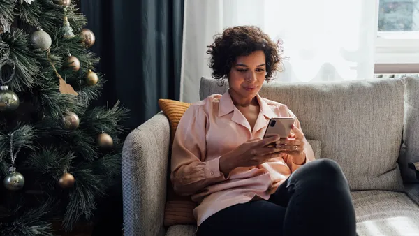 A person sits on a couch holding a mobile phone next to a Christmas tree.