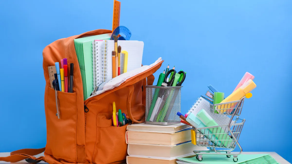 Orange backpack filed with school supplies and a toy-sized supermarket grocery cart with office supplies against a blue background.