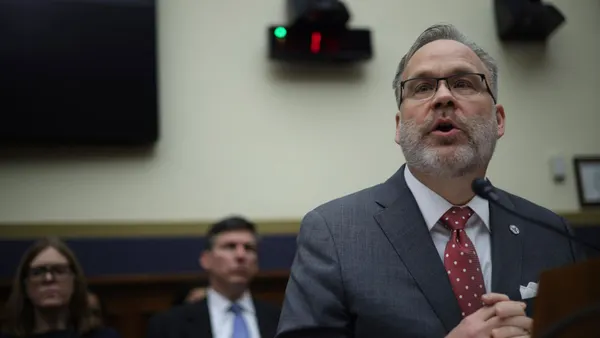Todd Harper speaks into a microphone while two people sit behind him; a screen and speakers are mounted on the wall above the people.
