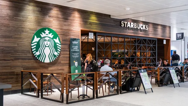 An image of a small Starbucks storefront with brown paneling and green siren logo inside an airport. Several people are sitting in chairs with luggage.
