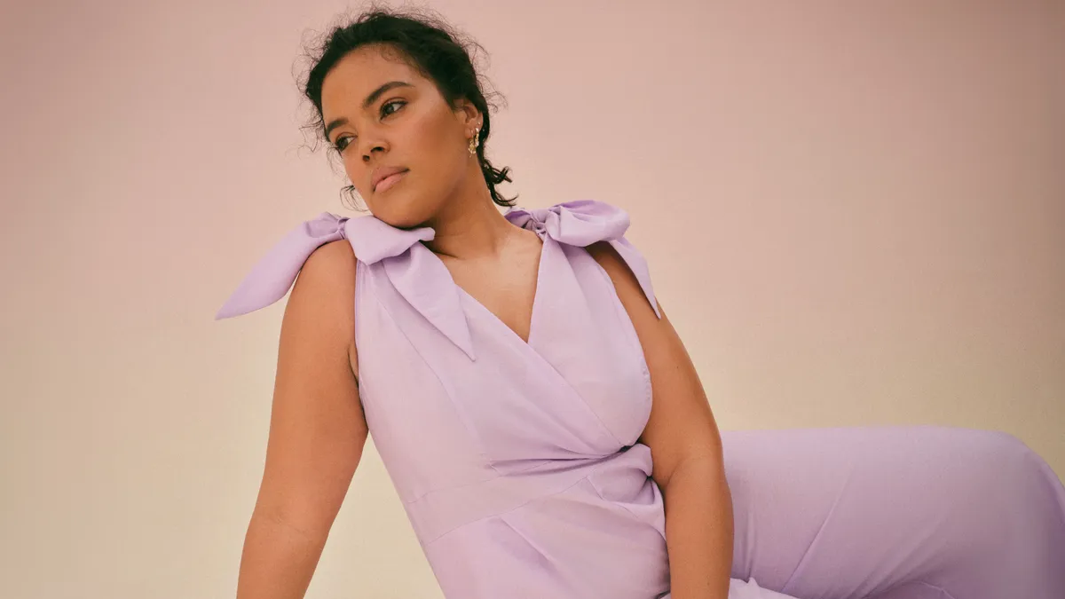 A woman in a lavender jumpsuit sits on one hip staring into the distance