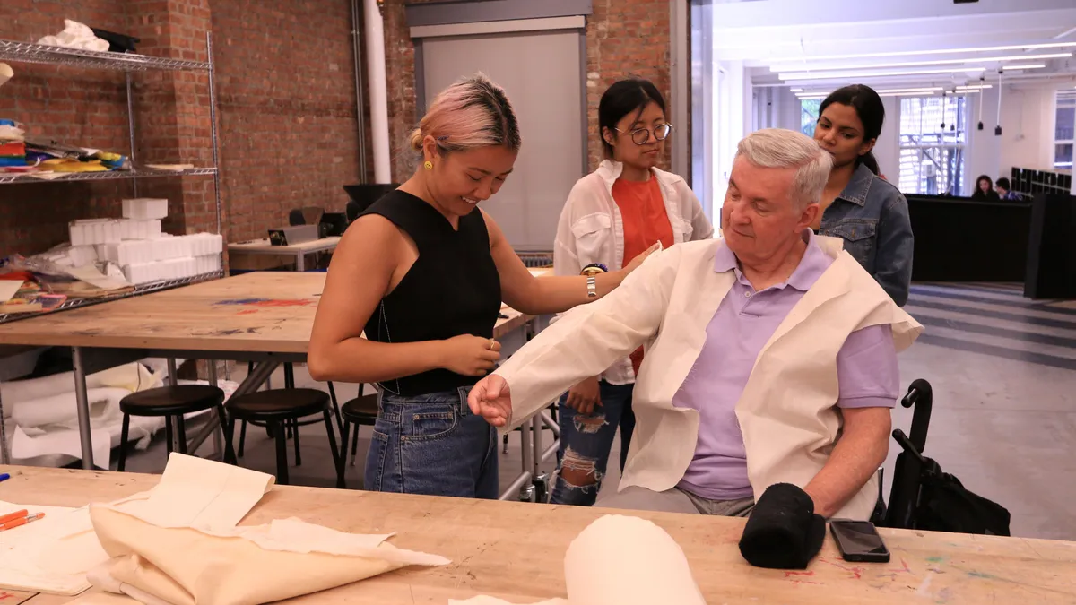 Behind a wide table with cut pieces of fabric, a person with pink hair a black top and blue denim jeans is standing next to a person with gray hair wearing a purple short sleeved collared shirt and sitting in a wheelchair. Two people watch from the background.