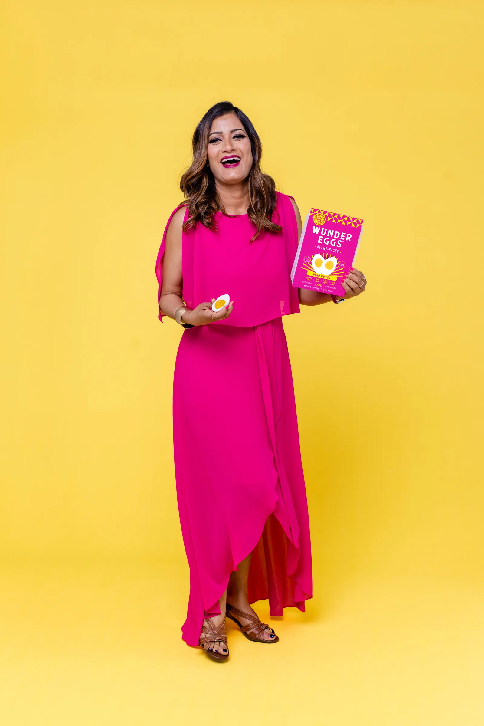 Hema Reddy smiles in a bright pink dress, holding a package of WunderEggs in her left hand and a WunderEgg half in her right. The background is yellow.
