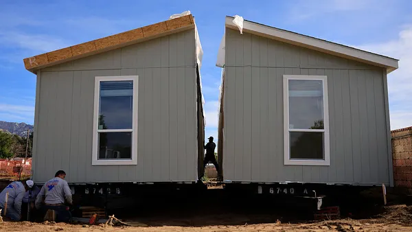 Construction workers assemble a squat prefabricated house with two windows under a blue sky.