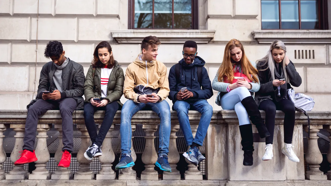 A row of six teenagers are sitting on a outdoor bench. Most are looking down at cellphones in their hands.