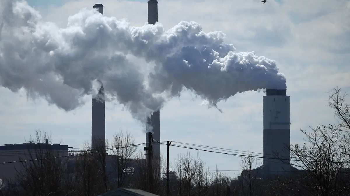 Heavy smoke clouds of carbon emissions spew from three cylindrical columns rising from a factory in the background, partly obscured by trees, with the roof of a house barely visible in the foreground.