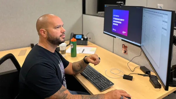 Tony Dotts works at his desk with two computer monitors. The screen on the left reads “Cybersecurity Awareness Month.”