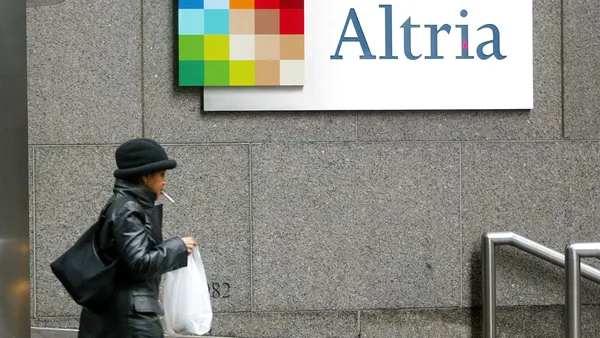 A photo of the exterior of a building, with a sign on the wall. The sign says "Altria." A person walks by the sign.