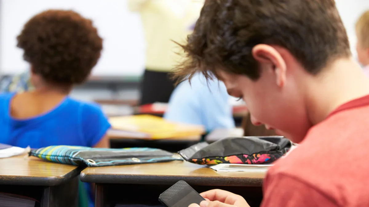 A student is sitting at a desk in a classroom holding a cellphone.