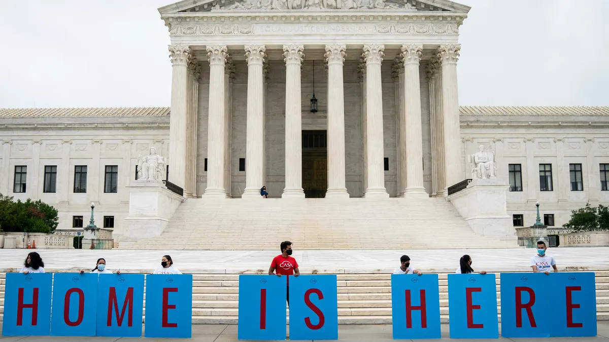 People stand in front of the Supreme Court with signs that read Home is Here.