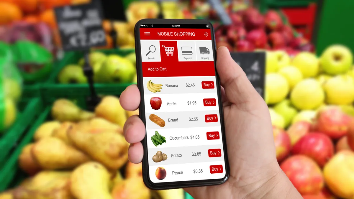 In front of a fruit display, a hand holds a phone with a grocery app open.