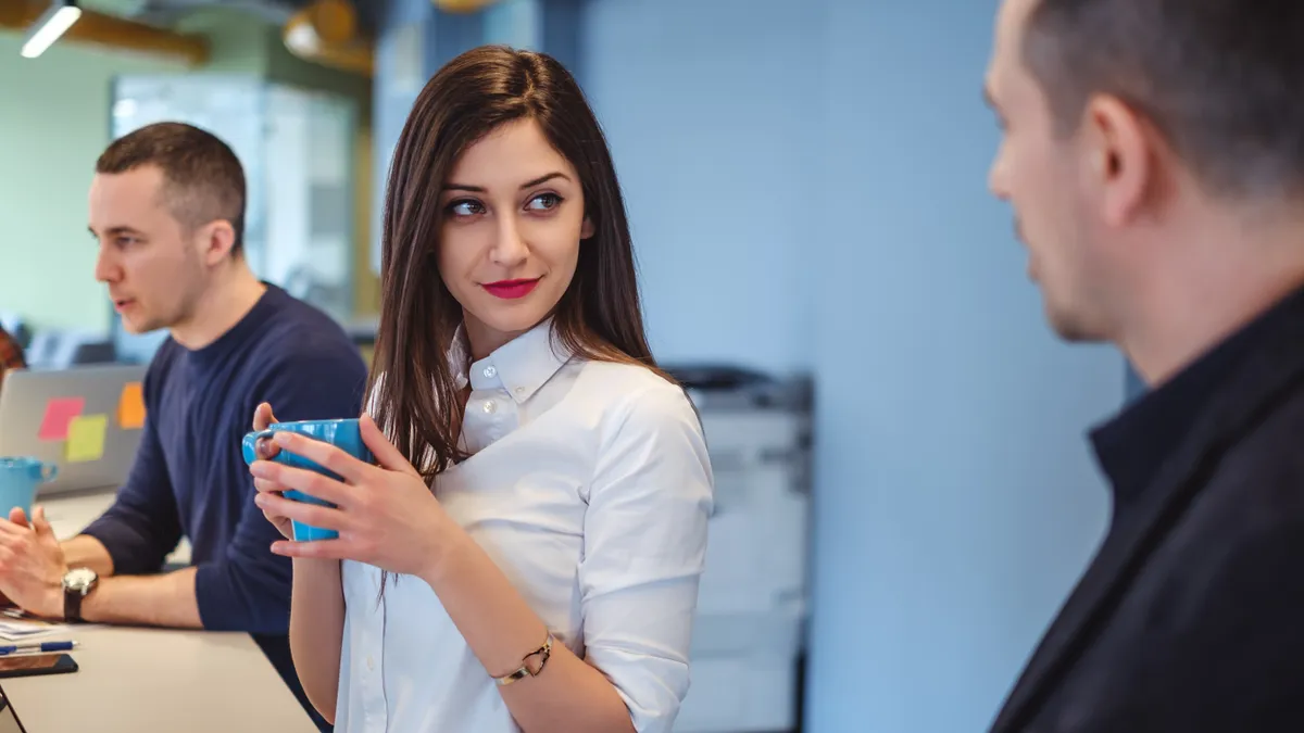 A woman looks at her colleague flirtatiously during a meeting while holding a mug.