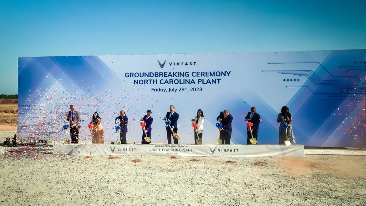 A collection of people in business attire stand against a backdrop curtain in the middle of a barren area. They have shovels. They are shoving the shovels into the dirt.