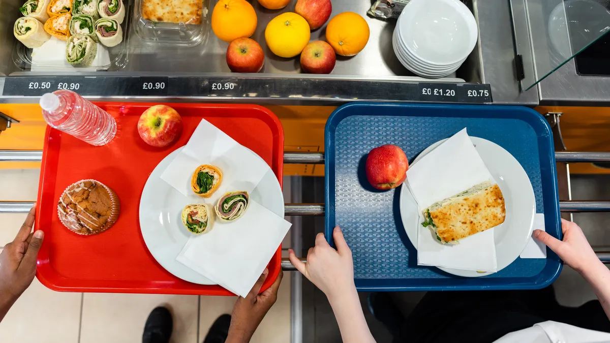 An overhead view of two students holding trays with sandwiches as they wait in line in a cafeteria.