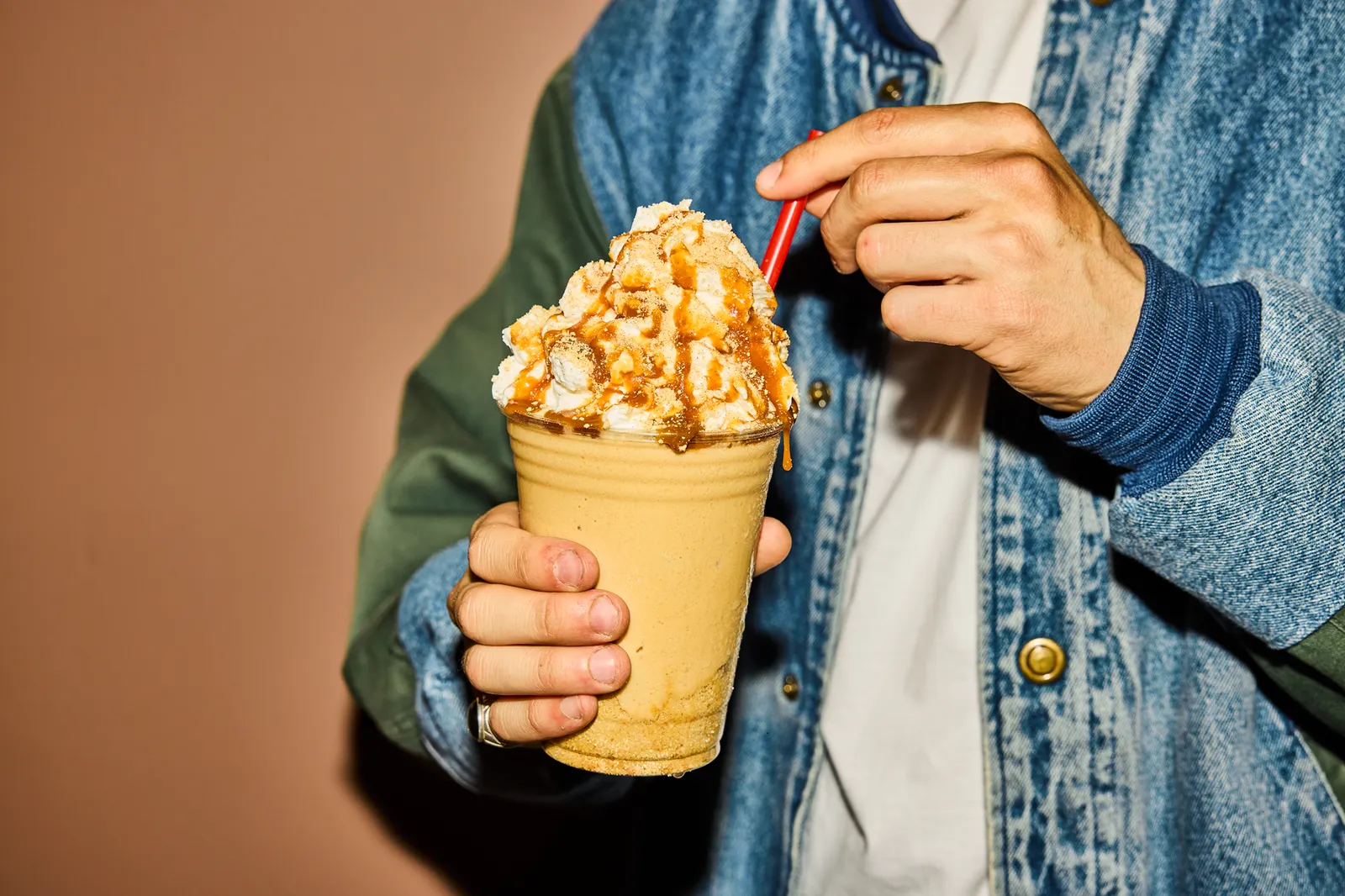 A photo of a person in a blue jacket standing in a room with brown walls, holding a milkshake.