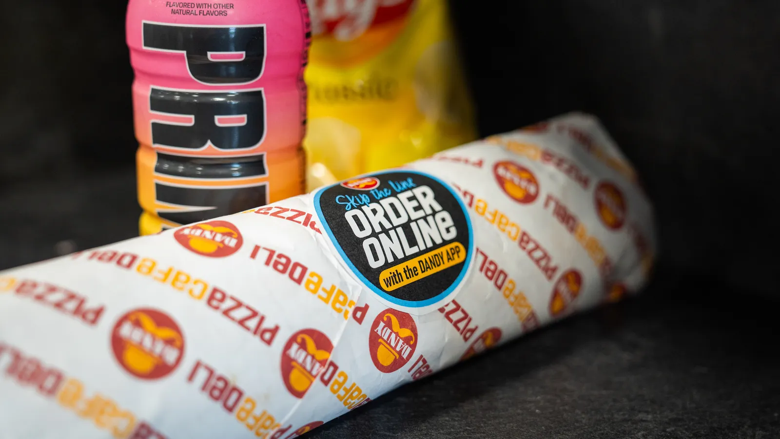 A photo of a bottled drink, a bag of Lays chips and a wrapped food item on a black background. The wrapped item has a sticker that says &quot;Skip the line, order online with the Dandy app.