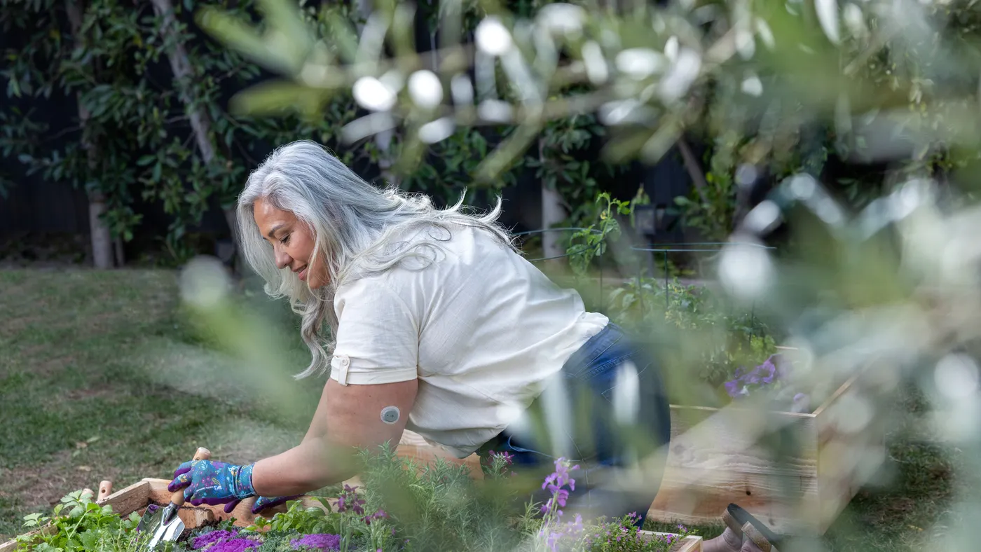 A person digs with a spade in a raised garden bed. They wear a gray sticker on their upper arm.