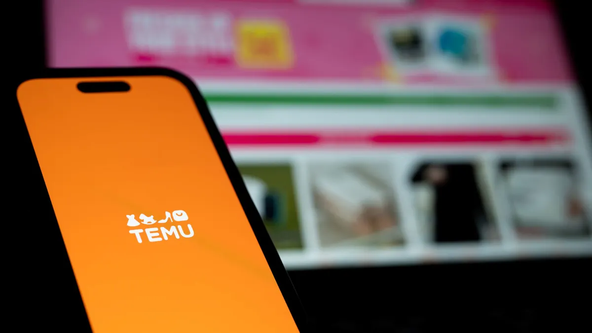 A phone displaying the Temu logo is seen in front of a computer screen.