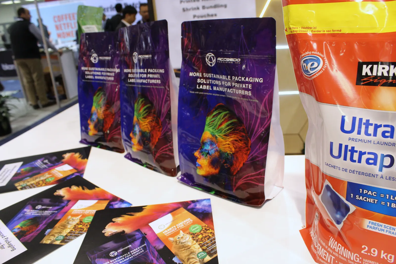 Various colorfully printed plastic pouches and pamphlets with Accredo Packaging branding sit on a trade show display.