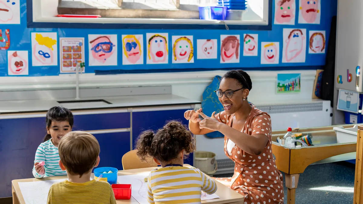 An adult is sitting at a small table with young students in a classroom. The adult is making a motion with their hands and the students are watching. Paintings of faces are on the back wall.