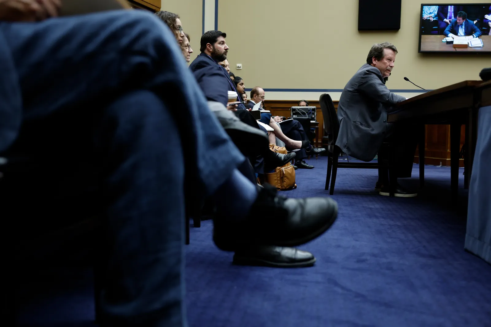 A person in a suit on the far right testifies to Congress into a microphone while a row of people sit behind watching. 